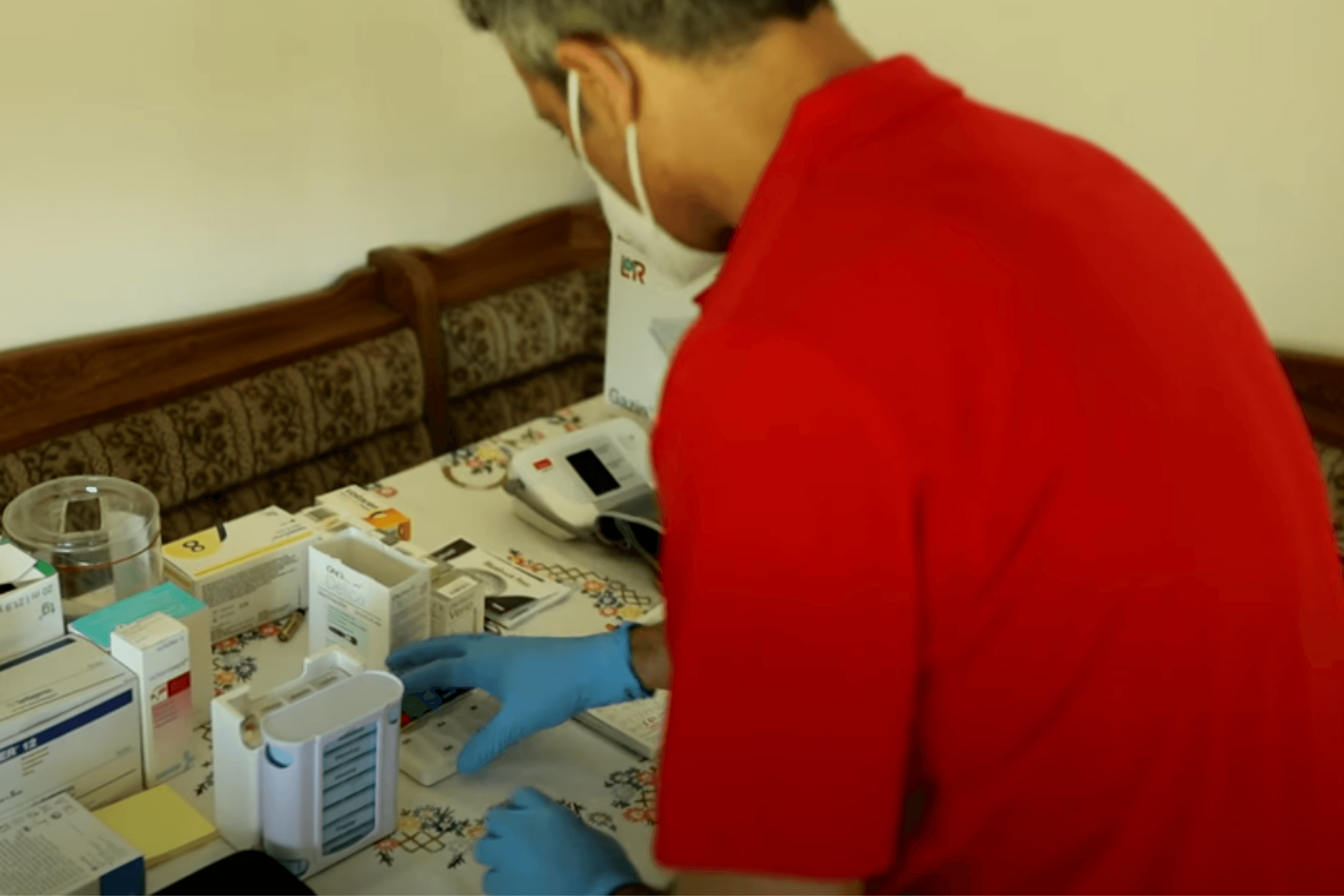 Ein Pflegeassistent mit einer Atemschutzmaske und blauen Gummihandschuhen der Volkshilfe Wien steht über einer Vielzahl an Medikamenten, die sich auf einem Tisch befinden. Er verschafft sich einen Überblick und sortiert diese.