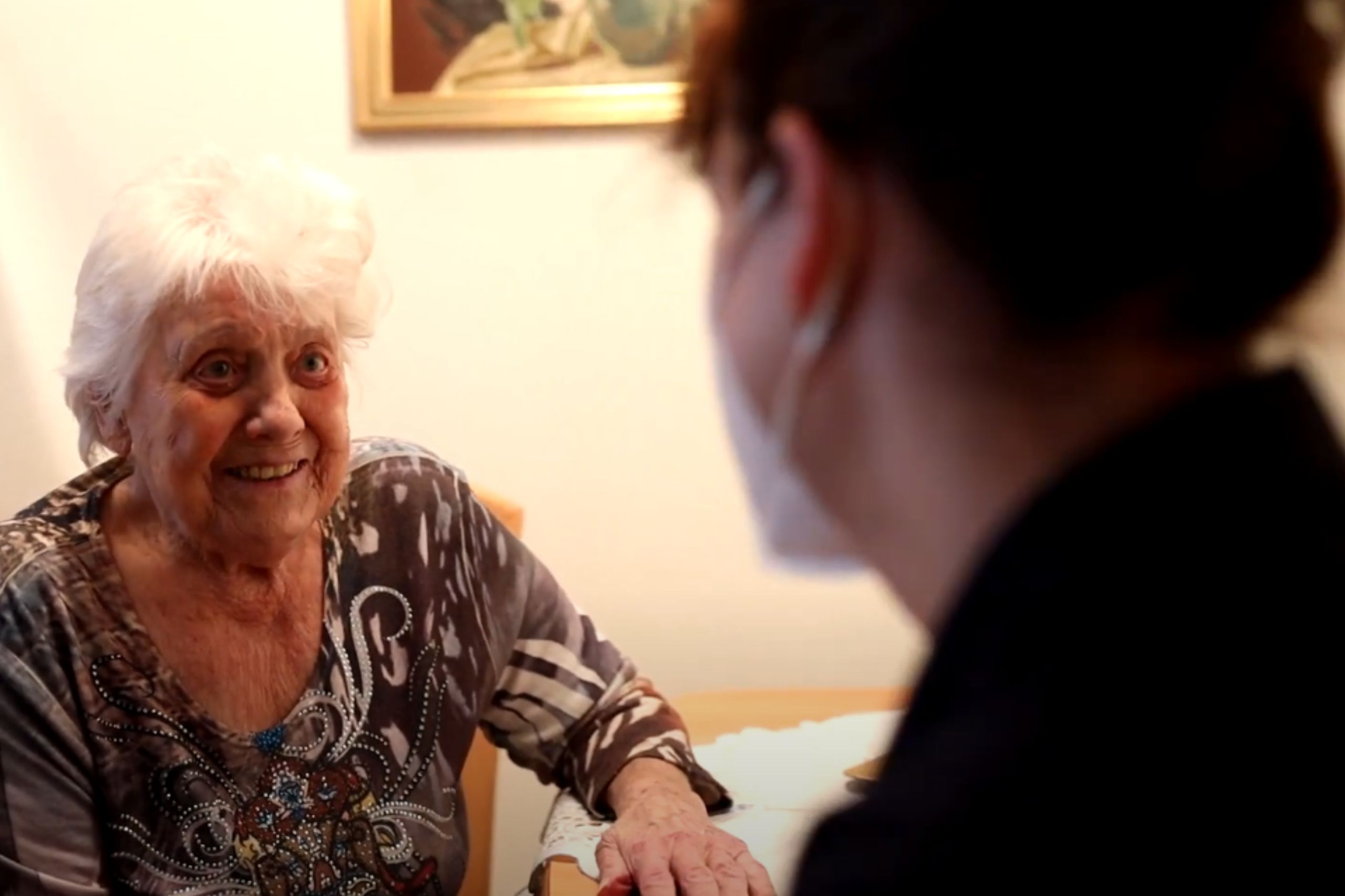 Links sitzt eine ältere Dame. Sie lächelt eine Heimhelferin der Volkshilfe Wien an, die rechts von ihr sitzt und eine Atemschutzmaske trägt. Beide sitzen an einem Tisch. Der Hintergrund ist ein Zimmer mit einem Gemälde.